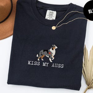 May include: Black t-shirt with the phrase "KISS MY AUSS" in white text. The shirt features an embroidered Australian Shepherd dog design with a red heart. A gold necklace and a brown hat are also visible.