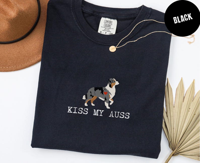 May include: Black t-shirt with the phrase "KISS MY AUSS" in white text. The shirt features an embroidered Australian Shepherd dog design with a red heart. A gold necklace and a brown hat are also visible.