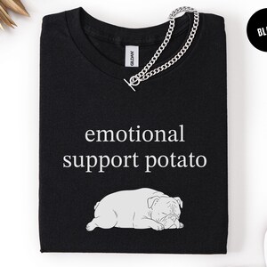 May include: Black t-shirt with the text "emotional support potato" and a graphic of a sleeping bulldog. A silver chain necklace is draped across the shirt. The word "Black" is in a black circle in the upper right corner.