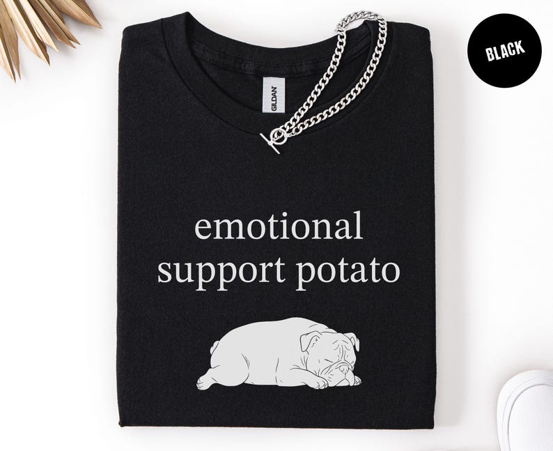 May include: Black t-shirt with the text "emotional support potato" and a graphic of a sleeping bulldog. A silver chain necklace is draped across the shirt. The word "Black" is in a black circle in the upper right corner.