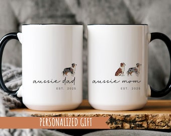 Könnte beinhalten: Zwei weiße Keramikbecher mit schwarzen Griffen und Rändern. Auf einem Becher steht "aussie dad" mit einer Hundeillustration und "EST. 2025". Der andere Becher trägt die Aufschrift "aussie mom" mit zwei Hundeillustrationen und "EST. 2025".