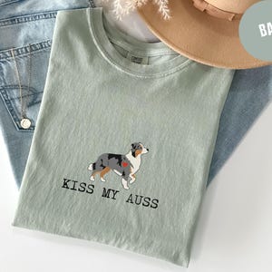 May include: A light green t-shirt with the text "KISS MY AUSS" and a graphic of an Australian Shepherd dog with a red heart. The shirt is folded on top of blue jeans, with a gold necklace and a tan hat.