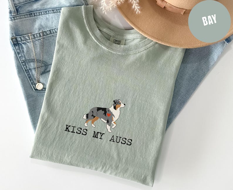 May include: A light green t-shirt with the text "KISS MY AUSS" and a graphic of an Australian Shepherd dog with a red heart. The shirt is folded on top of blue jeans, with a gold necklace and a tan hat.