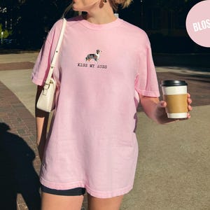 May include: A light pink t-shirt with the text "KISS MY AUSS" and a small dog graphic. The shirt is a loose fit and has short sleeves. A white purse is over the shoulder. A person is holding a coffee cup with a brown sleeve.