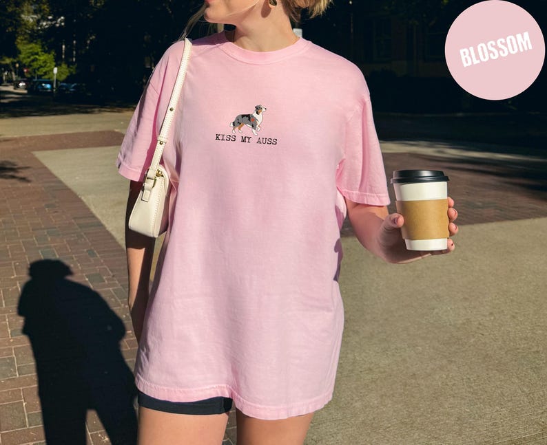 May include: A light pink t-shirt with the text "KISS MY AUSS" and a small dog graphic. The shirt is a loose fit and has short sleeves. A white purse is over the shoulder. A person is holding a coffee cup with a brown sleeve.
