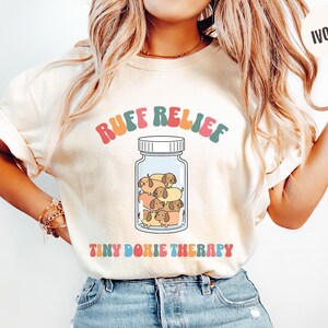 Puede incluir: Camiseta de color crema con la frase "Ruff Relief" en letras retro coloridas. Un gráfico de un tarro contiene perros salchicha de dibujos animados. Debajo del tarro, el texto "Tiny Doxie Therapy". La camiseta es de estilo casual y cómodo.