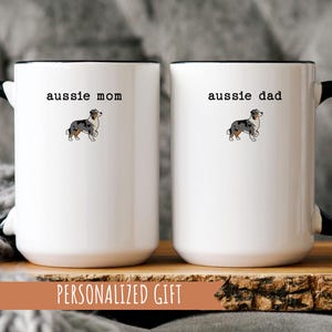 May include: Two white ceramic mugs with black handles and rims. One mug reads "aussie mom" and the other reads "aussie dad", each with a small illustration of an Australian Shepherd dog. The mugs are on a wooden surface.