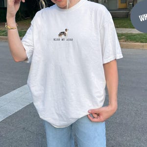 May include: White t-shirt with a small dog illustration and the text "KISS MY AUSS" in black. The shirt is a relaxed fit and has short sleeves. The dog illustration is in shades of brown, black, and white.