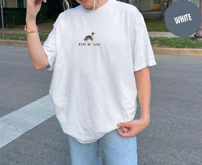 May include: White t-shirt with a small dog illustration and the text "KISS MY AUSS" in black. The shirt is a relaxed fit and has short sleeves. The dog illustration is in shades of brown, black, and white.