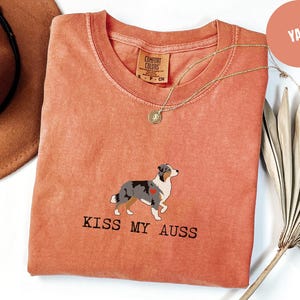 May include: A coral-colored t-shirt with a crew neck and the text "KISS MY AUSS" printed in black. The shirt features an illustration of an Australian Shepherd dog with a red heart. A gold necklace is draped on the shirt.