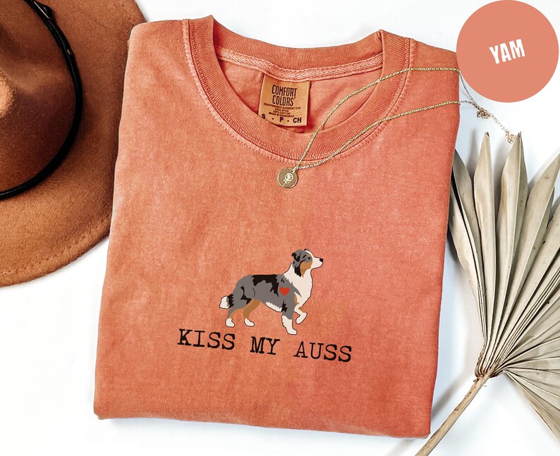 May include: A coral-colored t-shirt with a crew neck and the text "KISS MY AUSS" printed in black. The shirt features an illustration of an Australian Shepherd dog with a red heart. A gold necklace is draped on the shirt.