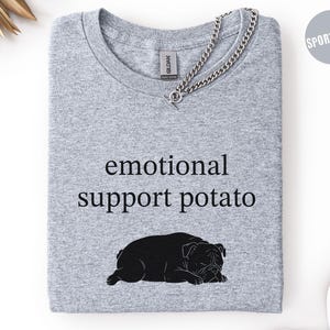 May include: A light gray t-shirt with the words "emotional support potato" in black, along with a black bulldog graphic. The shirt has a crew neck and a silver chain necklace. The shirt is folded neatly.