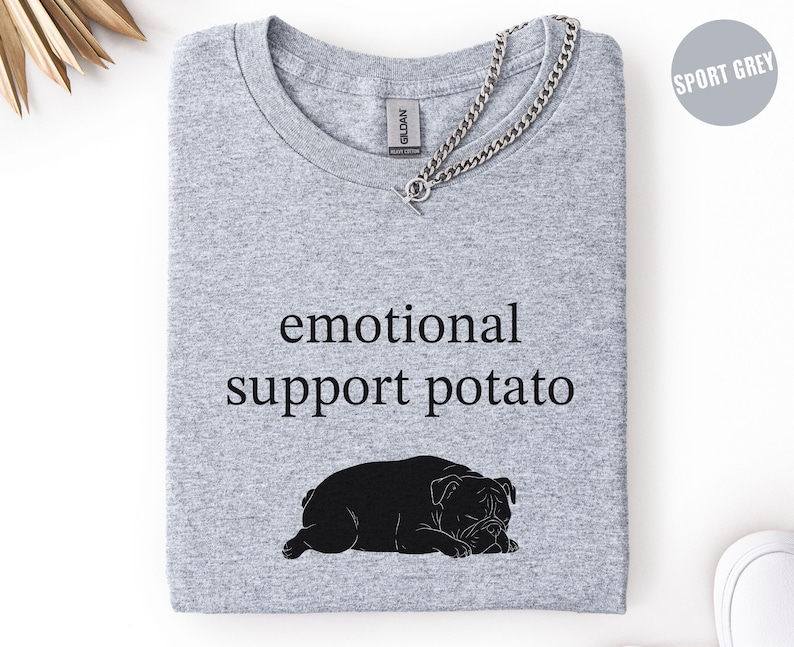 May include: A light gray t-shirt with the words "emotional support potato" in black, along with a black bulldog graphic. The shirt has a crew neck and a silver chain necklace. The shirt is folded neatly.