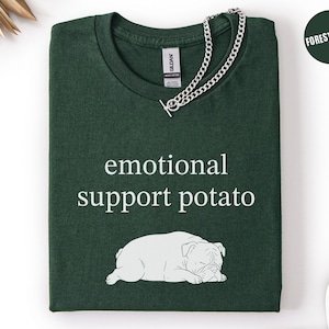 May include: Forest green t-shirt with the text "emotional support potato" and a white illustration of a sleeping bulldog. A silver chain necklace is draped over the shirt.