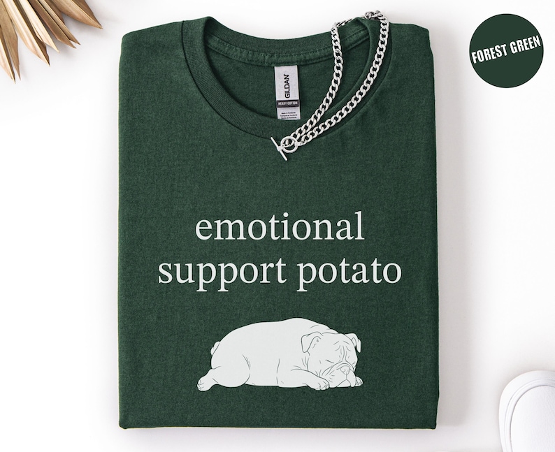May include: Forest green t-shirt with the text "emotional support potato" and a white illustration of a sleeping bulldog. A silver chain necklace is draped over the shirt.