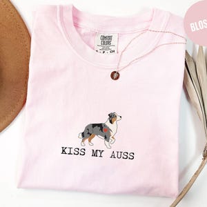 May include: A light pink t-shirt with a crew neck features a graphic of an Australian Shepherd dog with a heart and the text "KISS MY AUSS" in black. A gold necklace with a pendant is visible.