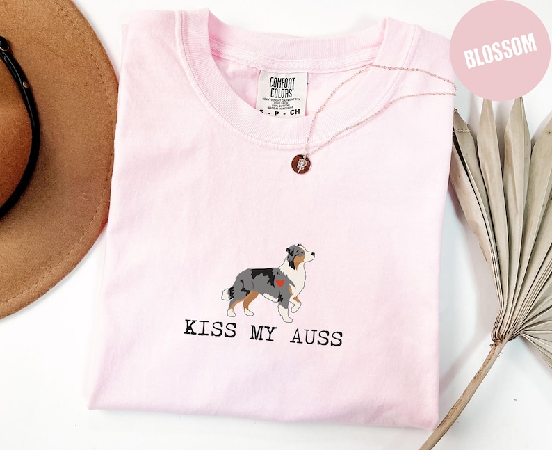 May include: A light pink t-shirt with a crew neck features a graphic of an Australian Shepherd dog with a heart and the text "KISS MY AUSS" in black. A gold necklace with a pendant is visible.