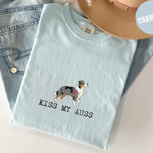 May include: Light blue t-shirt with a graphic of an Australian Shepherd dog and the text "KISS MY AUSS". The shirt is folded on top of a pair of blue jeans and a tan hat.
