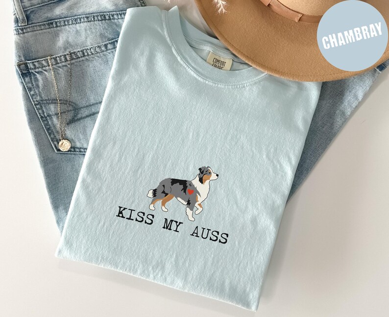 May include: Light blue t-shirt with a graphic of an Australian Shepherd dog and the text "KISS MY AUSS". The shirt is folded on top of a pair of blue jeans and a tan hat.