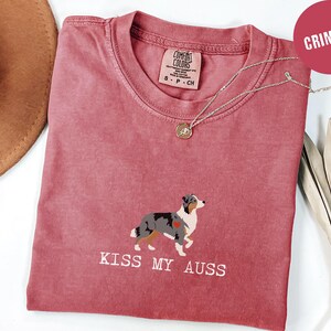 May include: Crimson-colored t-shirt with a graphic of an Australian Shepherd dog and the text "KISS MY AUSS". The shirt has a crew neck and short sleeves. A silver necklace with a pendant is draped across the shirt.