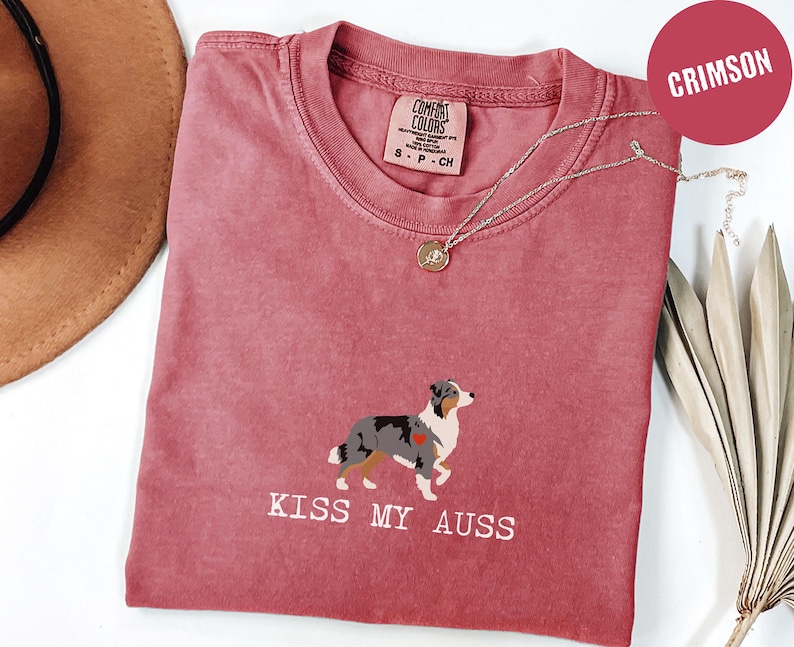May include: Crimson-colored t-shirt with a graphic of an Australian Shepherd dog and the text "KISS MY AUSS". The shirt has a crew neck and short sleeves. A silver necklace with a pendant is draped across the shirt.