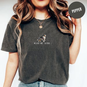 May include: Dark gray t-shirt with a small embroidered image of a dog and the text "KISS MY AUSS" in white. The dog is a mix of colors, including black, white, and brown. The shirt is a Pepper color.