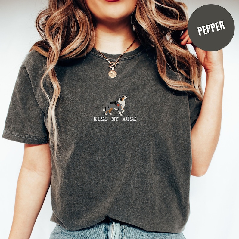 May include: Dark gray t-shirt with a small embroidered image of a dog and the text "KISS MY AUSS" in white. The dog is a mix of colors, including black, white, and brown. The shirt is a Pepper color.