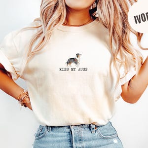 May include: Ivory-colored t-shirt featuring a graphic of an Australian Shepherd dog. The text "KISS MY AUSS" is printed below the dog illustration. The shirt is made of a soft material and has short sleeves.