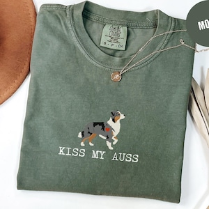 May include: A moss green t-shirt with a graphic of an Australian Shepherd dog and the text "KISS MY AUSS" in white. The shirt has a crew neck and short sleeves. A gold necklace is draped across the shirt.