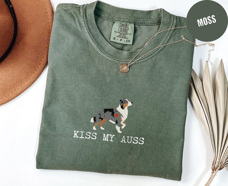 May include: A moss green t-shirt with a graphic of an Australian Shepherd dog and the text "KISS MY AUSS" in white. The shirt has a crew neck and short sleeves. A gold necklace is draped across the shirt.
