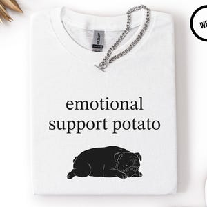 May include: White t-shirt with the text "emotional support potato" and a black illustration of a bulldog. A silver chain is draped across the shirt. The shirt is folded and displayed on a white surface.