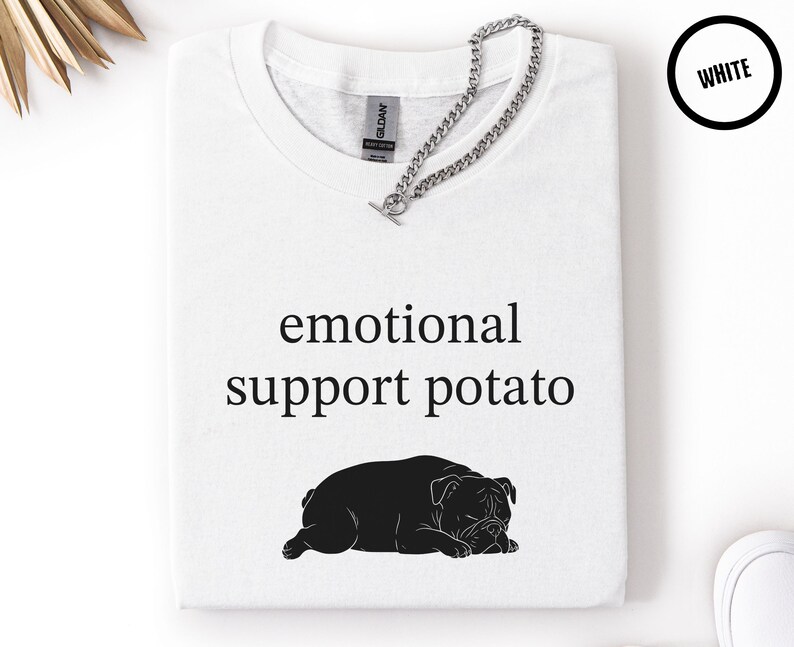 May include: White t-shirt with the text "emotional support potato" and a black illustration of a bulldog. A silver chain is draped across the shirt. The shirt is folded and displayed on a white surface.