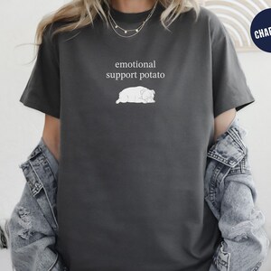 May include: Charcoal gray t-shirt with the text "emotional support potato" above a white illustration of a potato. The shirt has short sleeves and a relaxed fit. A navy blue circle with the word "CHARCOAL" is in the upper right corner.