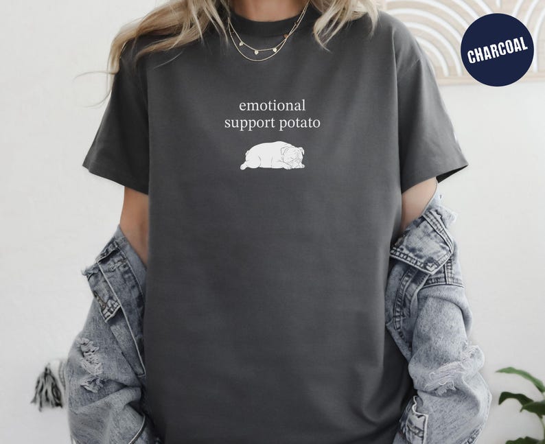 May include: Charcoal gray t-shirt with the text "emotional support potato" above a white illustration of a potato. The shirt has short sleeves and a relaxed fit. A navy blue circle with the word "CHARCOAL" is in the upper right corner.