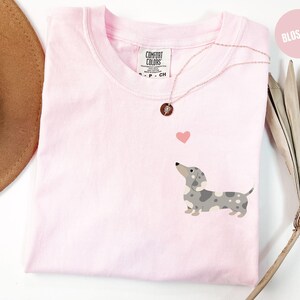 Peut inclure: T-shirt rose clair avec un motif de teckel et un petit cœur rose. Le t-shirt porte une étiquette Comfort Colors et un collier en or rose. Le mot "Blossom" est dans un cercle rose en haut à droite.