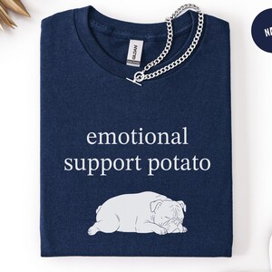 May include: Navy blue t-shirt with the text "emotional support potato" and a white illustration of a bulldog. A silver chain necklace is draped over the shirt. The shirt is folded neatly.