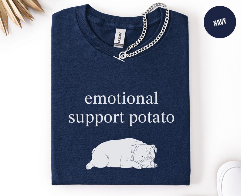 May include: Navy blue t-shirt with the text "emotional support potato" and a white illustration of a bulldog. A silver chain necklace is draped over the shirt. The shirt is folded neatly.