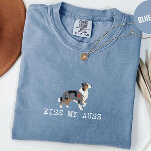May include: Blue jean-colored t-shirt with a crew neck. The shirt features an embroidered Australian Shepherd dog with a red heart and the text "KISS MY AUSS" in white. A gold necklace with a pendant is draped on the shirt.