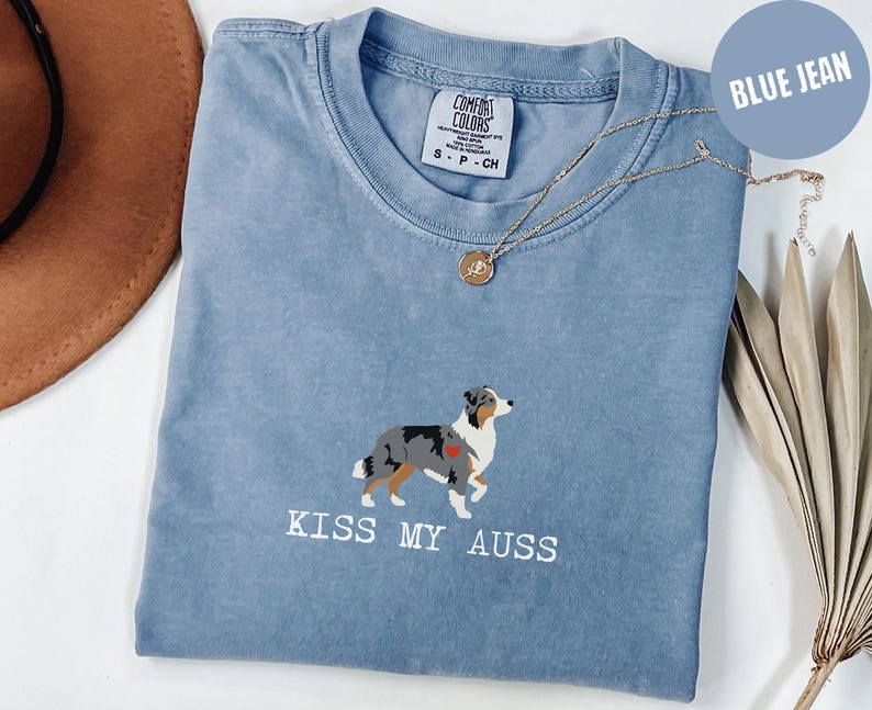May include: Blue jean-colored t-shirt with a crew neck. The shirt features an embroidered Australian Shepherd dog with a red heart and the text "KISS MY AUSS" in white. A gold necklace with a pendant is draped on the shirt.