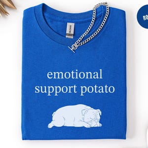 May include: Royal blue t-shirt with the text "emotional support potato" and a white bulldog graphic. A silver chain necklace is draped across the shirt. The shirt is folded and ready to wear.