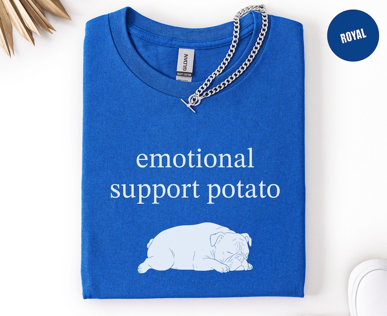 May include: Royal blue t-shirt with the text "emotional support potato" and a white bulldog graphic. A silver chain necklace is draped across the shirt. The shirt is folded and ready to wear.