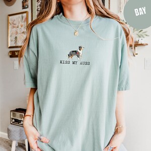 May include: A light teal t-shirt featuring a graphic of an Australian Shepherd dog with a red heart. The text "KISS MY AUSS" is printed below the dog. The shirt is a casual, unisex style.