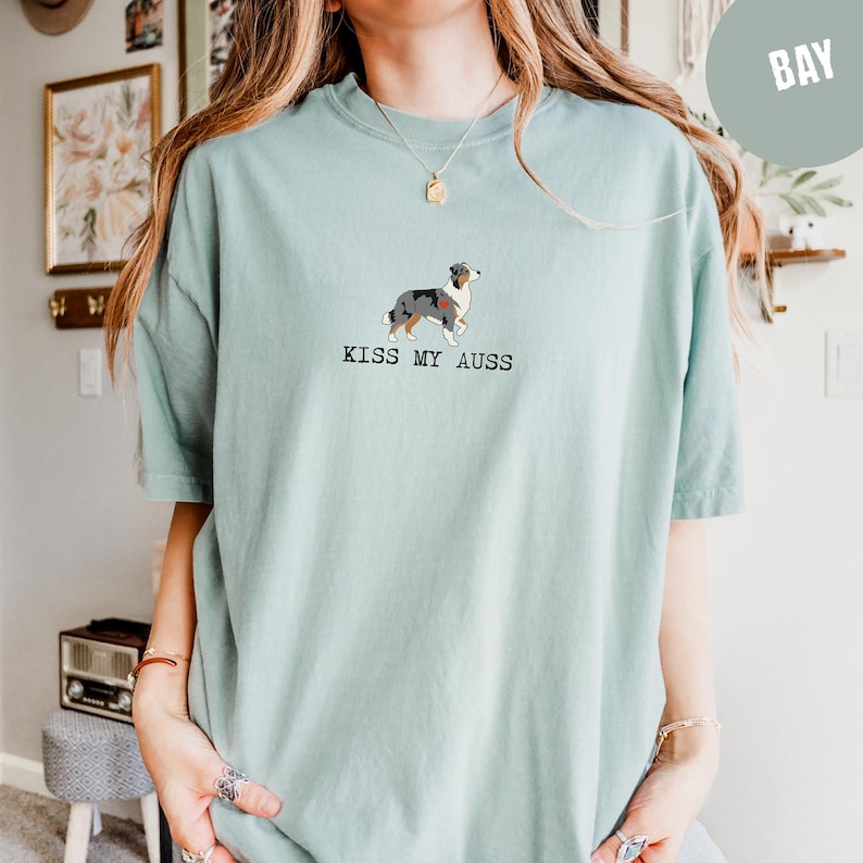 May include: A light teal t-shirt featuring a graphic of an Australian Shepherd dog with a red heart. The text "KISS MY AUSS" is printed below the dog. The shirt is a casual, unisex style.