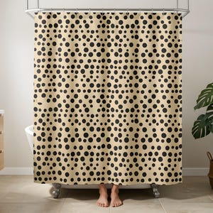 May include: A shower curtain featuring a repeating pattern of solid black circles of varying sizes set against a light beige background. The curtain hangs from a metal rod with rings and covers the length of a bathtub, displaying a consistent polka dot design.