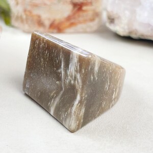 Pode incluir: Um anel triangular feito de pedra listrada de marrom e branco. O anel está sobre uma superfície branca.