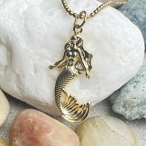 May include: A gold-tone necklace with a mermaid pendant. The mermaid is depicted with flowing hair and a tail.