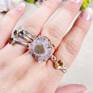 Pode incluir: Uma mão usando várias alianças, incluindo uma aliança de ouro com uma pedra de ametista roxa engastada em forma de flor.