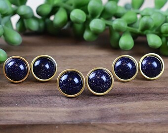 May include: Six pairs of round stud earrings with a dark blue, speckled appearance, set in gold-colored bezels. The earrings are arranged on a wooden surface, with green foliage in the background.