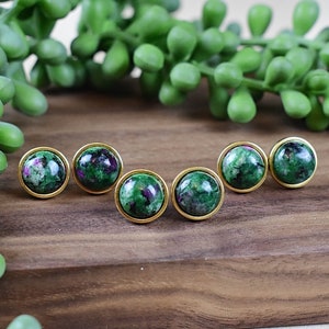 Pode incluir: Seis brincos redondos com moldura dourada. Os brincos apresentam uma pedra verde com inclusões vermelhas e pretas. Os brincos são exibidos em uma superfície de madeira com folhagem verde ao fundo.