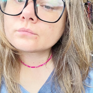 May include: A close-up of a person wearing a pink beaded choker necklace. The necklace is made of small, round beads that are strung together on a thin wire. The person is wearing a blue shirt and has long, blonde hair.